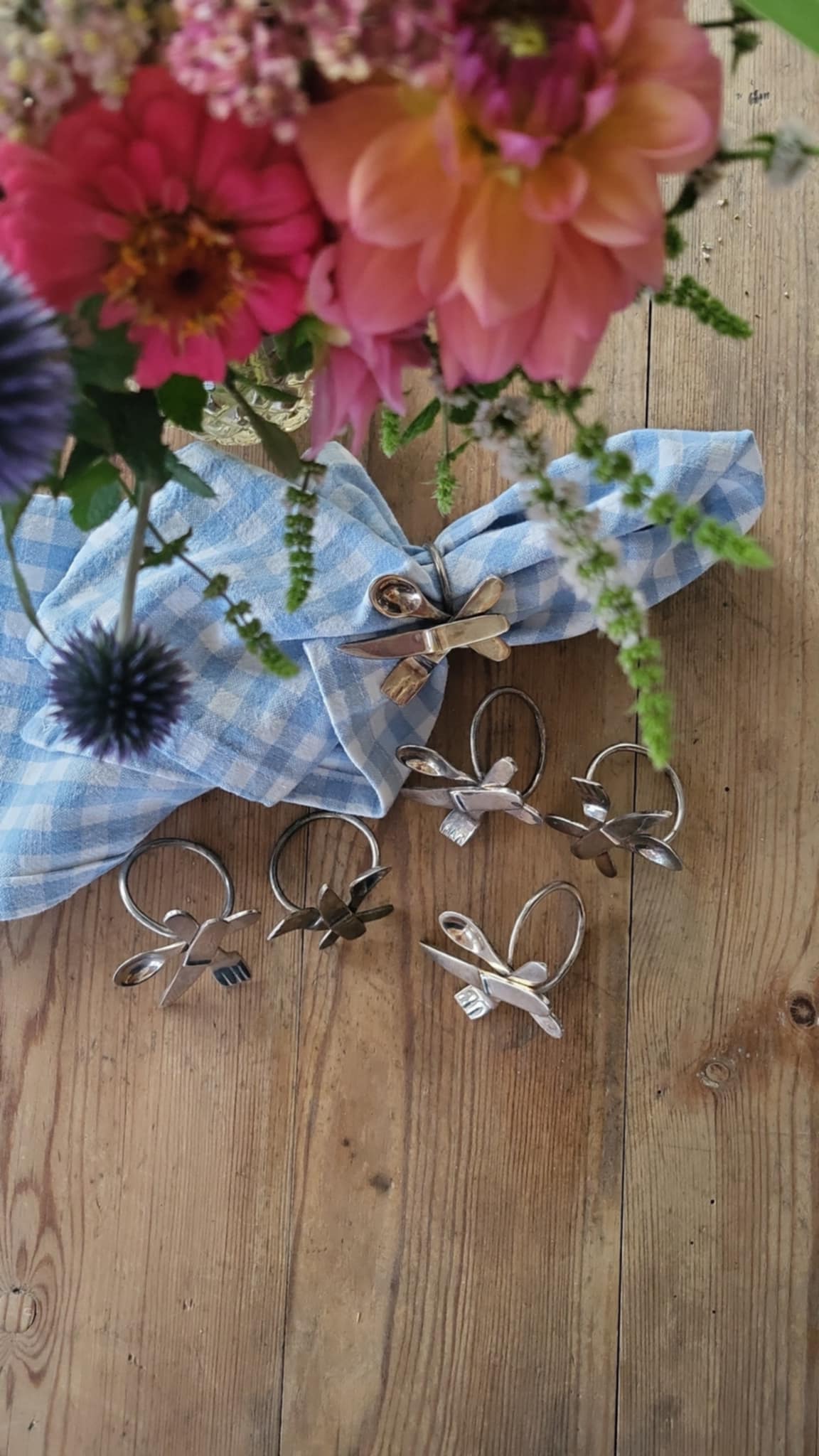 Servettringar i metall arrangerade på rustikt bord med linneservett och blommor i bakgrunden