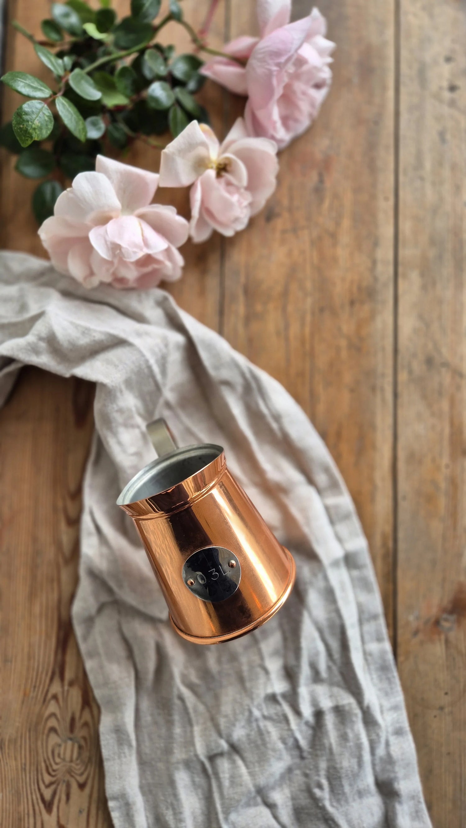 Ett decilitermått i koppar ligger på en linnéhandduk på ett träbord. I bakgrunden syns en bukett rosa blommor.