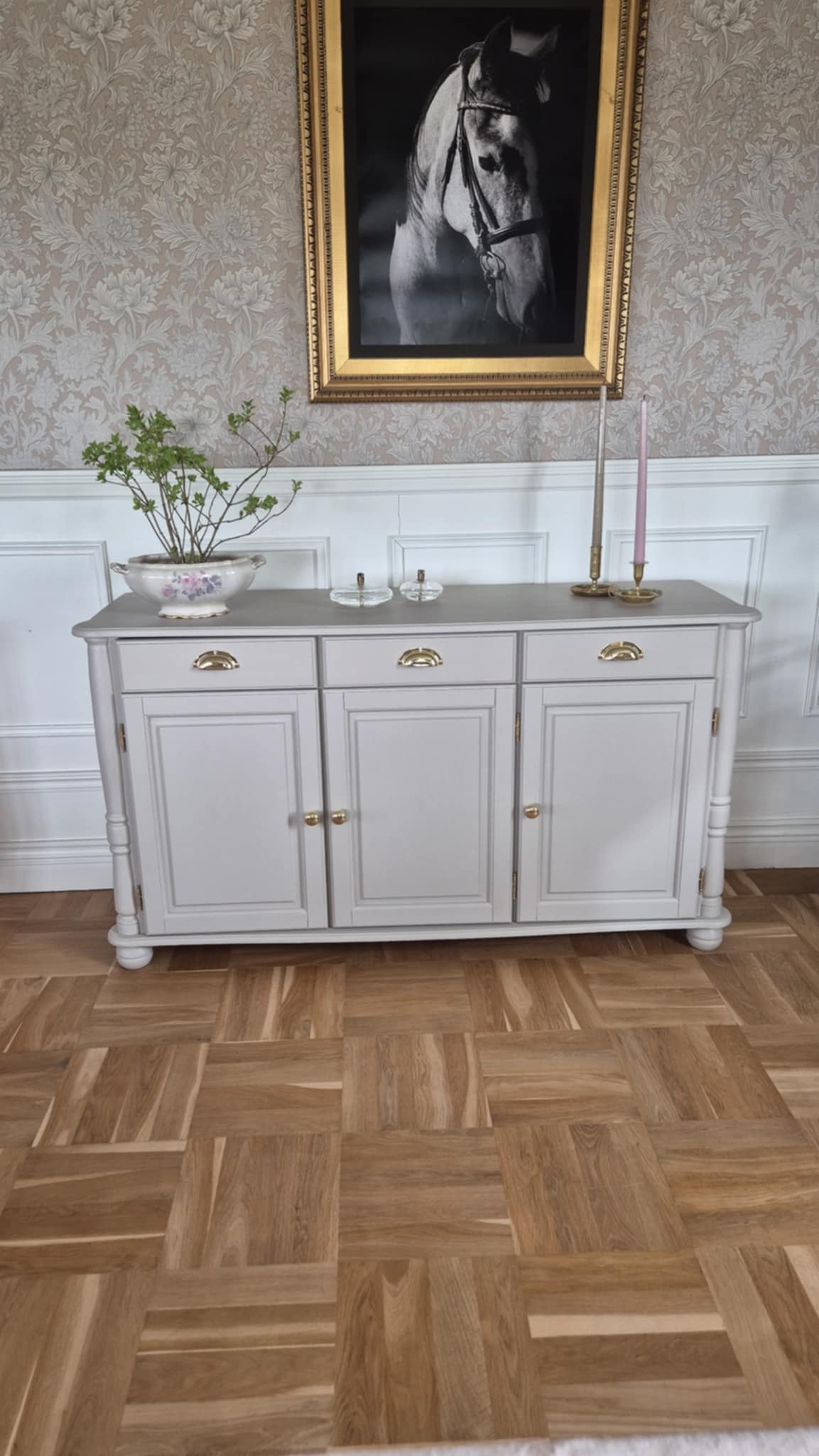 Renoverad skänk i ljusgrå/vit färg med tre lådor och tre skåpdörrar, guldiga skålhandtag och runda svarvade ben. Skänken står mot en vägg med paneler och blommigt tapet, på parkettgolv i fiskbensmönster.