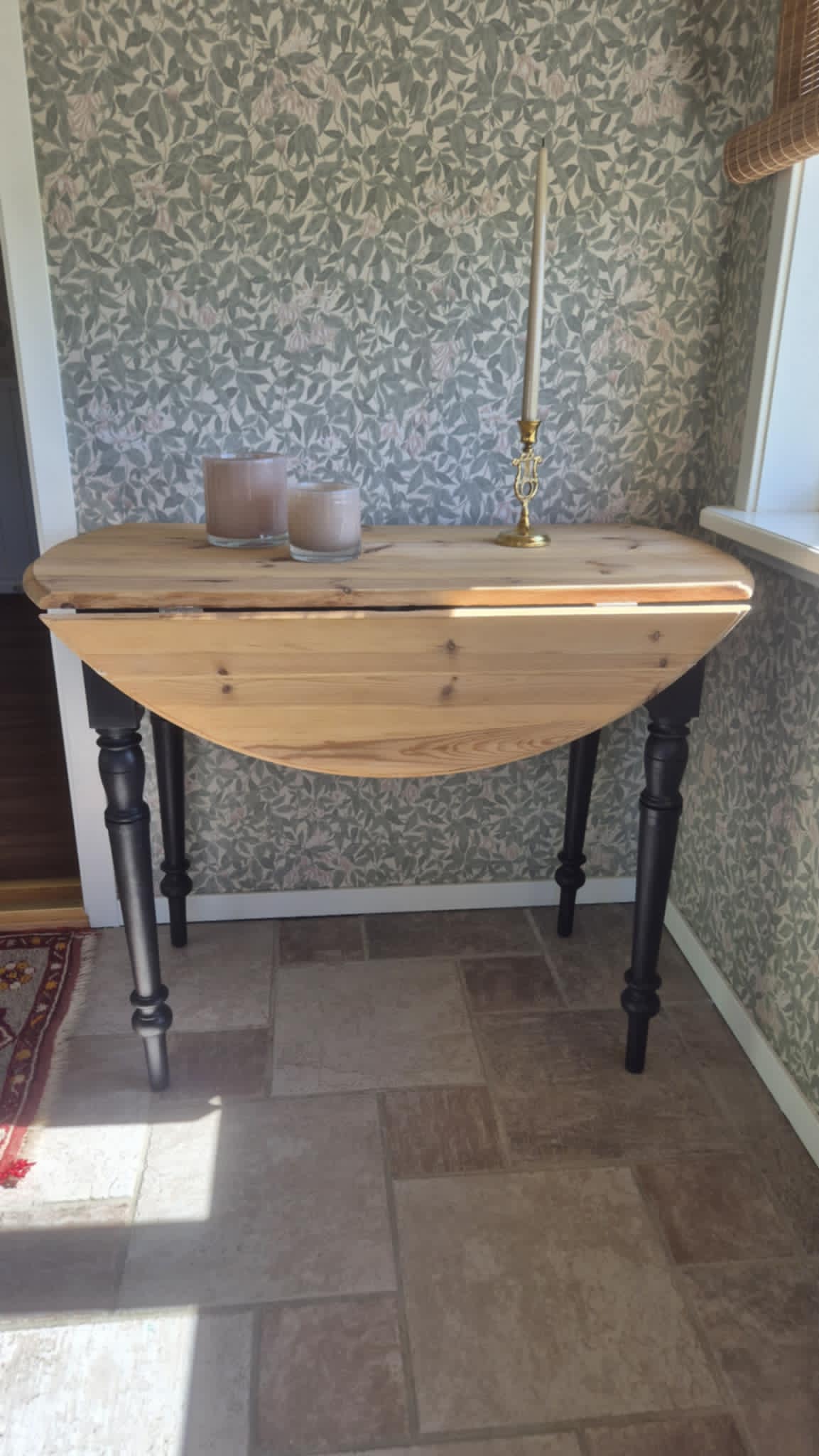 Runt slagbord med naturlig ljus träskiva som visar kvistar och årsringar, samt svartmålade klassiskt svarvade ben. Bordet har två utfällbara klaffar och står på ett stengolv mot en vägg med blommigt mönstrat tapet i gröna och beiga toner. På bordet står ljuslyktor och en guldlampa.